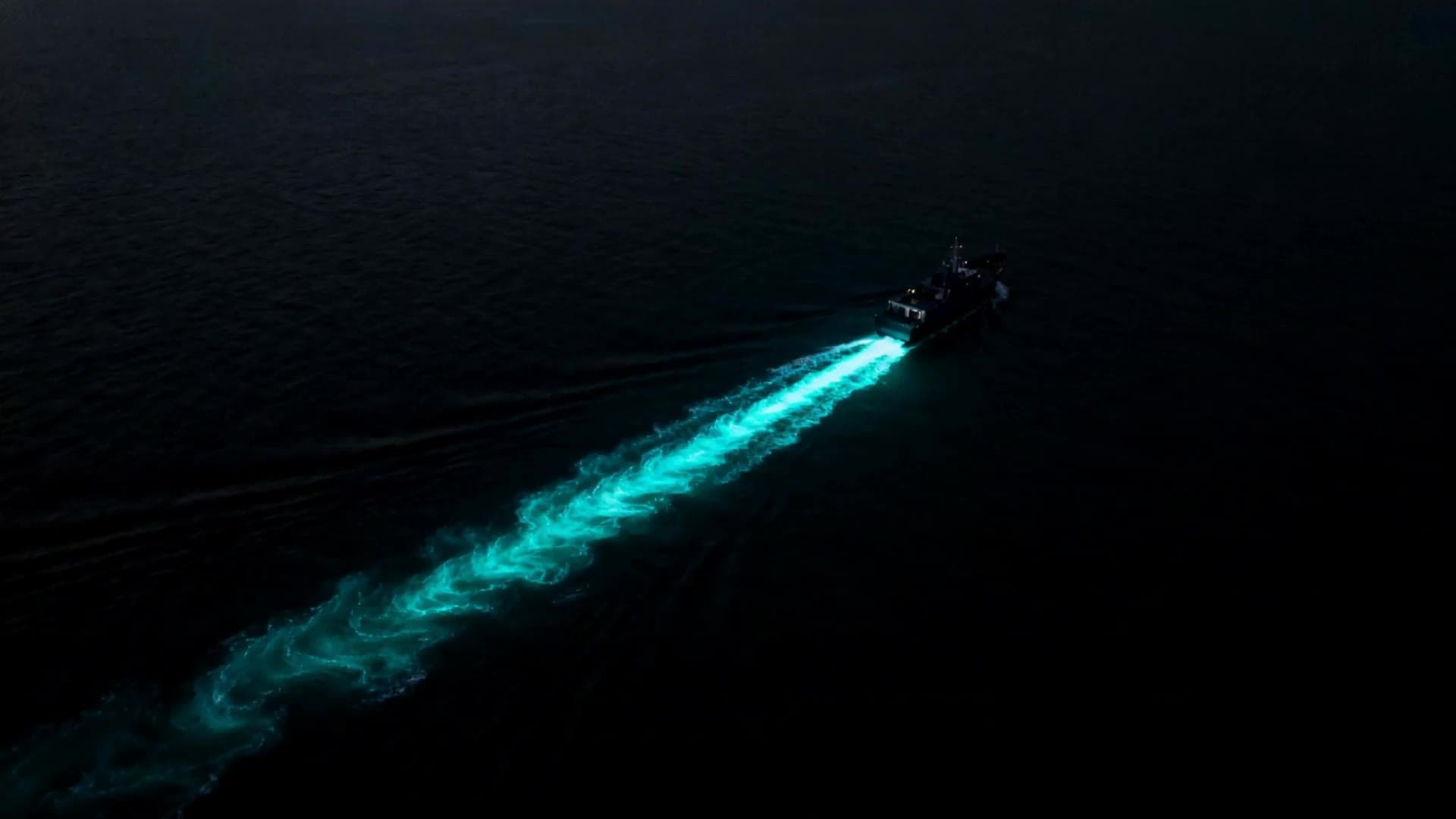 Aerial view of a dark vessel crossing a black ocean at night, its churning wake glowing bioluminescent teal
