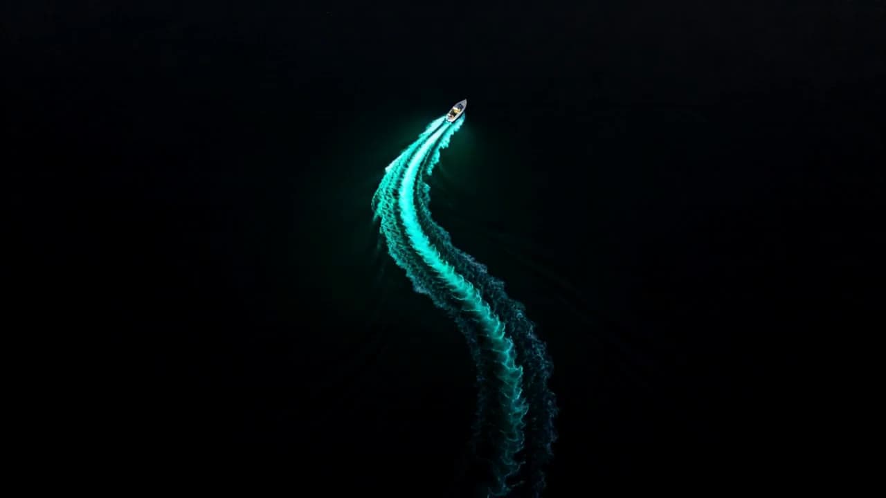 Top-down aerial of a small boat tracing an S-curved glowing teal wake across black water